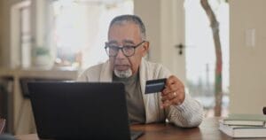 Persona de mediana edad revisando tarjeta de crédito con laptop en casa, preocupado por tasas de interés altas