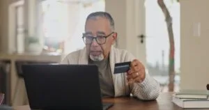 Persona de mediana edad revisando tarjeta de crédito con laptop en casa, preocupado por tasas de interés altas