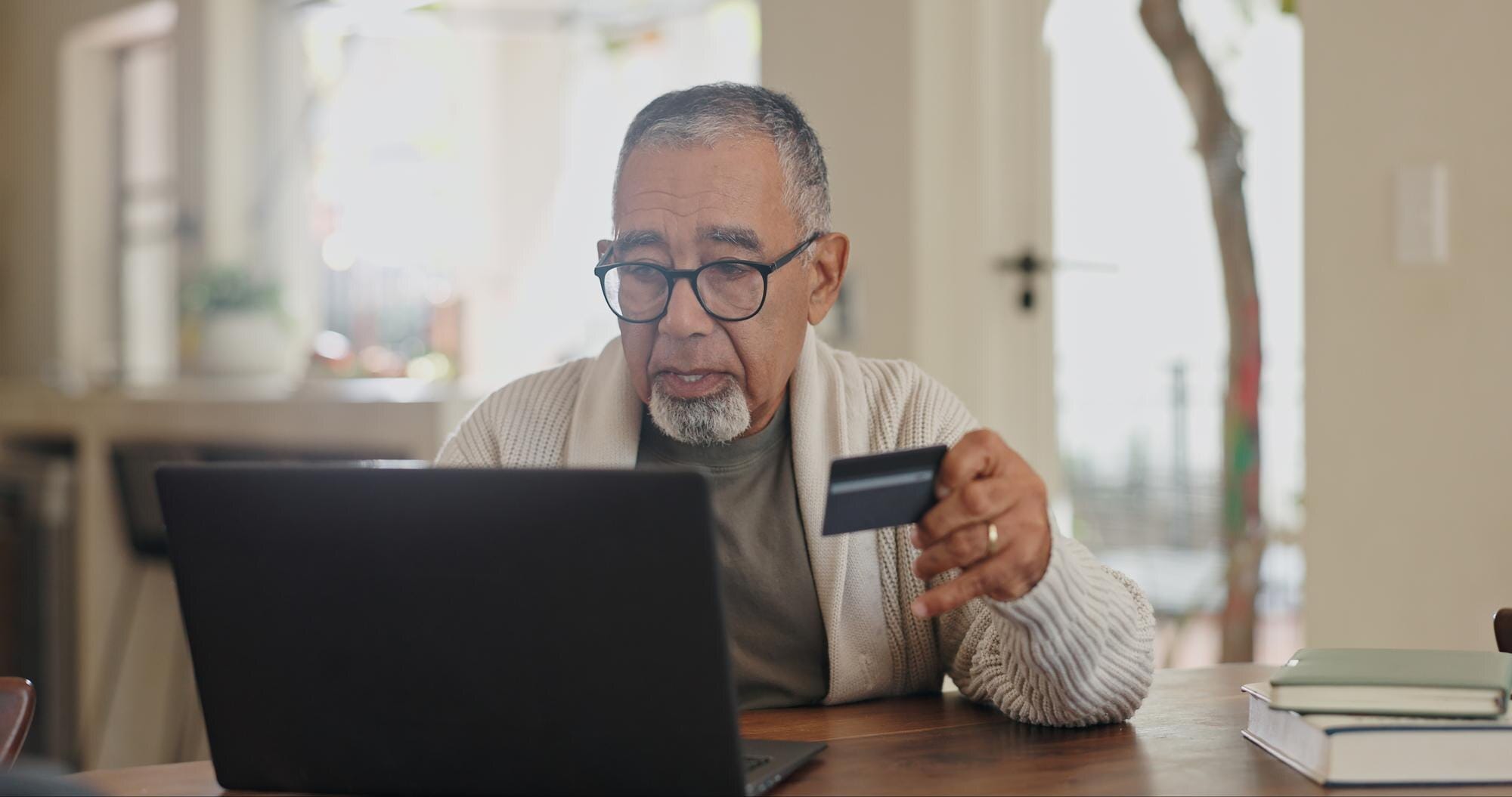 Persona de mediana edad revisando tarjeta de crédito con laptop en casa, preocupado por tasas de interés altas